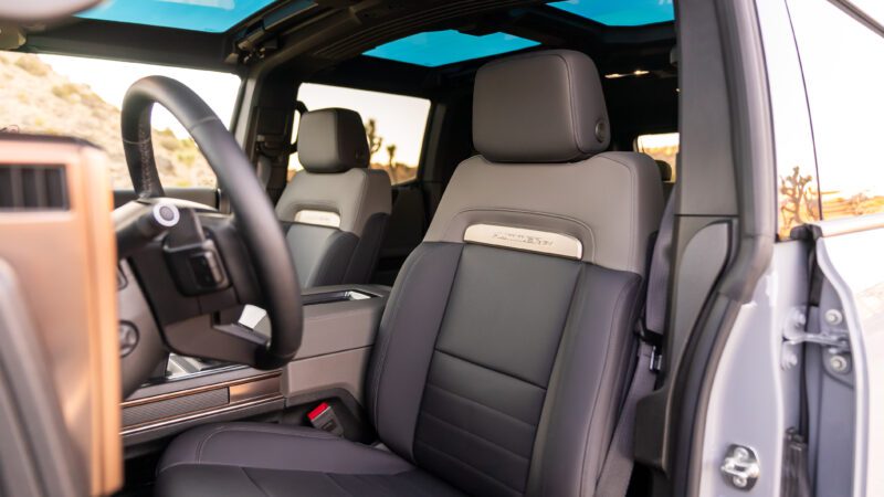 The interior view of this sleek EV SUV showcases black and gray leather front seats, a steering wheel, and a dashboard with elegant wood accents. Experience tranquility with the sky visible through the sunroof, offering true solace on every journey.